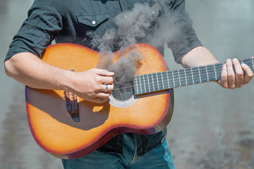a man in a jeans plays the guitar from which smoke comes, against the background of nature