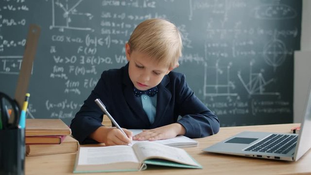 Prodigy kid in suit is taking notes sitting at desk in university classroom with chalkboard covered with formulas in background. Modern education and talanted children concept.