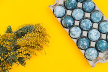 Blue textured colored Easter eggs in a box and mimosa flowers on a bright yellow holiday background backdrop. Spring holiday concept. Copyspace. Flat lay. Top View.