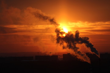 Red sun at sunset or dawn at dusk low above the horizon shrouded in smoke from the chimneys of the plant