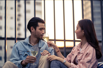 Asian young couple having celebrate anniversary party together on rooftop. Man and woman holding glass of white wine toasting and drinking with happiness and smile. Love of young boy and girl concept.