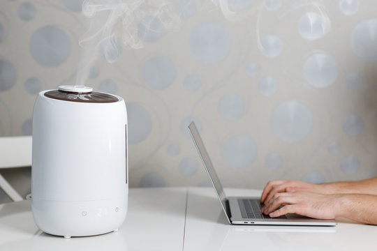 Air humidifier stay on the table and spreading steam. Humidification of the dry air. Comfortable living conditions. Near hands on laptop. Selective focus on vapor. Air purity and health care concept.