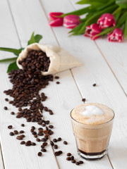 Cappuccino or latte with milk froth in a glass. Сoffee beans in a bag of coffee and coffee leaves on a white wooden background. Spring flowers, tulips in the background. Soft focus, close up.