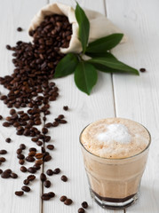 Cappuccino or latte with milk froth in a glass. Сoffee beans in a bag of coffee and coffee leaves on a white wooden background. Soft focus, close up.