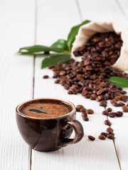 Close-up of dark coffee cup with espresso, roasted coffee beans in the bag of burlap, on white wooden background. Near coffee leaves. soft focus.