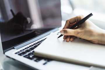 Woman writes with a pen in notebook on laptop keyboard in a sunny office, business and education...