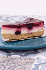 Slice of summer fruit cheesecake with a biscuit base served with a cake cutter utensil on a blue plate. Blue patterned tiled background