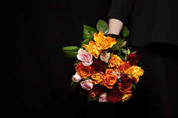 Bouquet of different color roses in female hand on a black background. Red, orange and pink flowers with water drops on petals. Romantic Valentine's Day Gift
