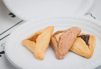 Hamantaschen. (ozen haman). Jewish food for Purim. Triangular crispy dough, stuffed with dates, halva and chocolate, on a white plate.
