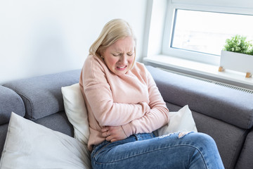 View of young woman suffering from stomachache on sofa at home. Woman sitting on bed and having stomach ache. Young woman suffering from abdominal pain while sitting on sofa at home