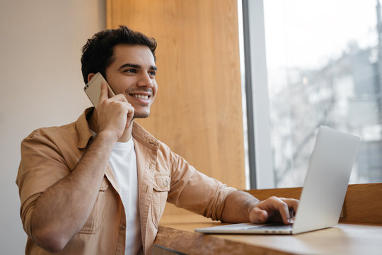 Indian Businessman With Happy Emotional Face Talking On Cell Phone, Sitting In Office. Successful Asian Man Planning Start Up, Using Laptop Computer And Mobile. Portrait Of Freelancer At Workplace