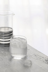 Glass of clear cold water with ice and crystal pot on white concrete background.