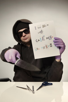 Criminal in hooded shirt preparing ransom letter from newspaper