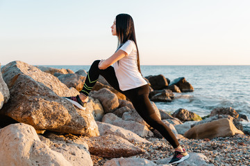 The concept of sport. A young brunette woman is engaged in stretching on the coastal rocks. In the background, the sea and the shore. In profile view