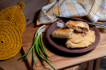 Clay plate of fried meat pies with cutlery and green onion on wooden table.