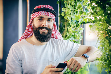 Young handsome trendy muscular bearded arab guy standing next to window in cafe, holding smart phone and looking at camera.
