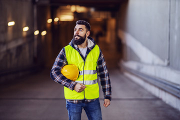 Young hardworking positive bearded construction worker in vest holding safety helmet under armpit...