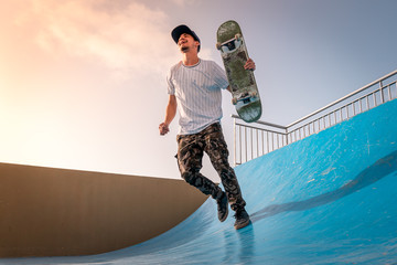 joven skater corre sonriendo a lo largo de la rampa de skateboard con el tablero en la mano al atardecer