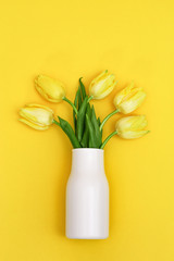 Blooming tulip flowers in small white vase on yellow background. Bouquet of flowers as spring gift  for women or mothers day. View from above and vertical format.
