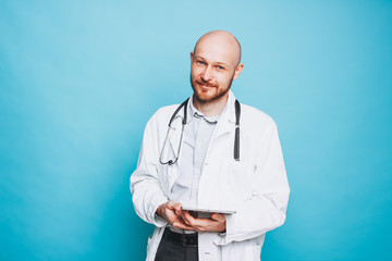 Attractive friendly bald bearded smiling doctor with tablet looking at camera isolated on blue background