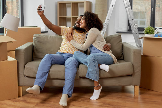 moving, repair and real estate concept - happy african american couple with cardboard boxes sitting on sofa and taking selfie by smartphone at new home