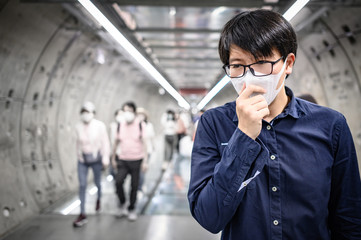 Asian man wearing surgical face mask coughing in subway station with crowded people walking pass. Wuhan coronavirus (COVID-19) outbreak prevention in public area. Health care and medical concept