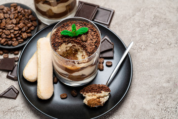 Classic tiramisu dessert in a glass cup, pieces of chocolate and savoiardi cookies on concrete background