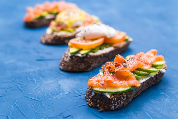 Sandwiches with smoked salmon, eggs, sauce and avocado on blue background. Concept of breakfast and healthy nutrition.
