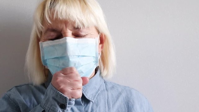 Adult 40 Year Old Blonde Girl In A Blue Shirt And A Medical Mask On The Background Of A Gray Wall Of The Hospital. Coronavirus, Colds And Pneumonia Concept