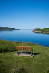 Obraz premium A bench overlooking a body of water, which is surrounded by grassy cliffs and hills, that drains out to see on a clear, cloudless day on the Isle of Skye, Scotland. 