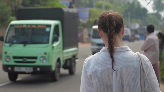 resting girl with braid and handbag walks along road past moving vehicles in tropical town backside view slow motion. Concept heavy traffic traveling