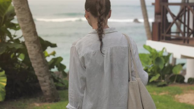 young woman walks near rest hotel against tropical plants and storming ocean backside view slow motion. Concept exotic travel environment