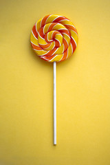 Bright swirl lollipop on yellow background. Festive vertical background with colorful twisted candy. Close up of rainbow round sweet on stick. Orange and yellow confectionery. Minimalistic design