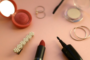 Beauty products and fashionable accessories on pale pink background: blush, mascara, eyeliner, highlighter, lipstick, beret, rings and hoop earrings. Selective focus.