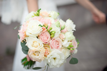 Close-up groom in a suit and the bride in a white dress are standing and holding a bouquet of peach roses, eustomas and flowers and greenery.