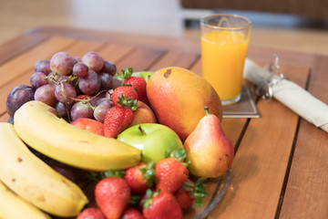 Fruits (bananas, apples, mango, grapes, strawberries) placed on a table next to a glass of orange juice ready for breakfast
