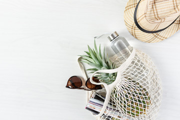 Mesh shopping bag with pineapple, glossy magazine and eco friendly bottle of water on white wooden background.