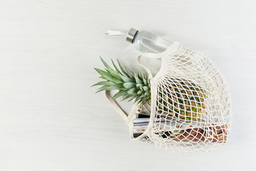 Mesh shopping bag with pineapple, glossy magazine and eco friendly bottle of water on white wooden background.