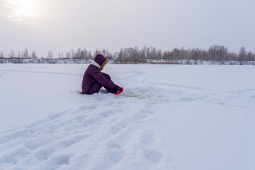 Fototapeta na wymiar Lonely girl angler fishing with a winter fishing rod sitting on a substrate on the ice among the white snow.