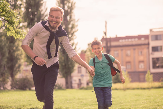 two people, young man and his son or younger brother, running in the grass field in the city. Looking at camera, excited and happy.