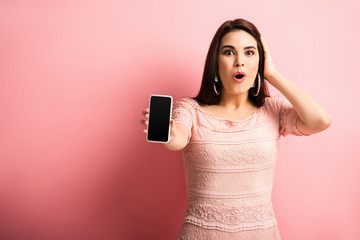 impressed girl touching head while showing smartphone with blank screen on pink background