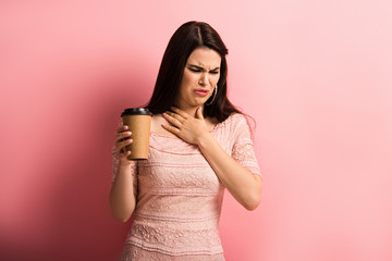 displeased girl touching chest while holding coffee to go on pink background