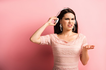 Fototapeta premium displeased girl touching wireless headphones while standing with open arm on pink background