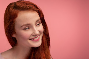 Fototapeta premium Portrait of smiling redhead girl. Perfect skin. Beautiful female model with long hair over pink background. Young cute woman with clear skin. Skincare and facial treatment concept.