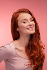 Fototapeta premium Portrait of smiling redhead girl. Perfect skin. Beautiful female model with long hair over pink background. Young cute woman with clear skin. Skincare and facial treatment concept.