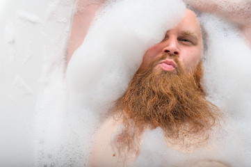 Portrait of a bald man with a long red beard takes a bath with foam. Top view on a cheerful funny...