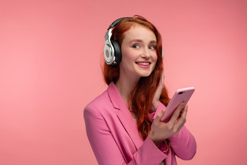 Enjoy listening to music. Beautiful young redhead woman with headphones listening music on smart phone using music app. Funny smiling girl with earphones and mobile phone on pink background.