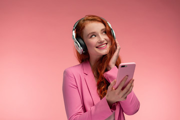 Enjoy listening to music. Beautiful young redhead woman with headphones listening music on smart phone using music app. Funny smiling girl with earphones and mobile phone on pink background.