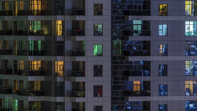 Rows of glowing windows with people in the interior of apartment building at night. Modern skyscraper from glass and concrete. Concept for business and modern life. Zoom out