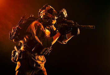 Army special operations soldier, commando fighter in full tactical ammunition, helmet with radio headset and night vision device, aiming short barrel assault rifle in darkness, low key studio shoot